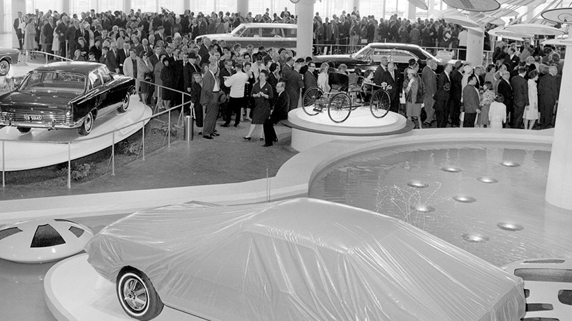 A black and white photo of four cars on separate display platforms with one under a cover all in front of a large crowd