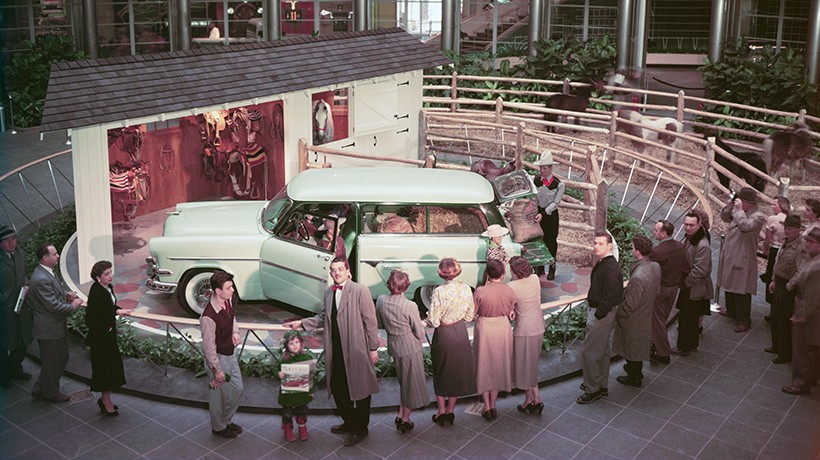 A small group of people observing an exhibit of a teal Ranch Wagon car