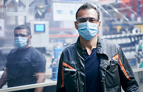 two men wearing face masks with machinery in shop