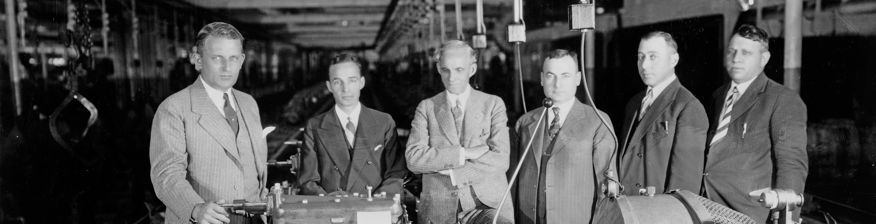A black and white photo of Henry Ford lined up with executives inspecting a Model A engine