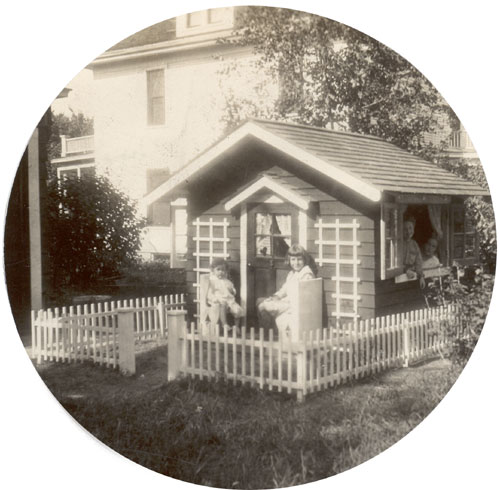 Image of a fenced in playhouse with two children on the porch and at least two people inside looking out of the window.